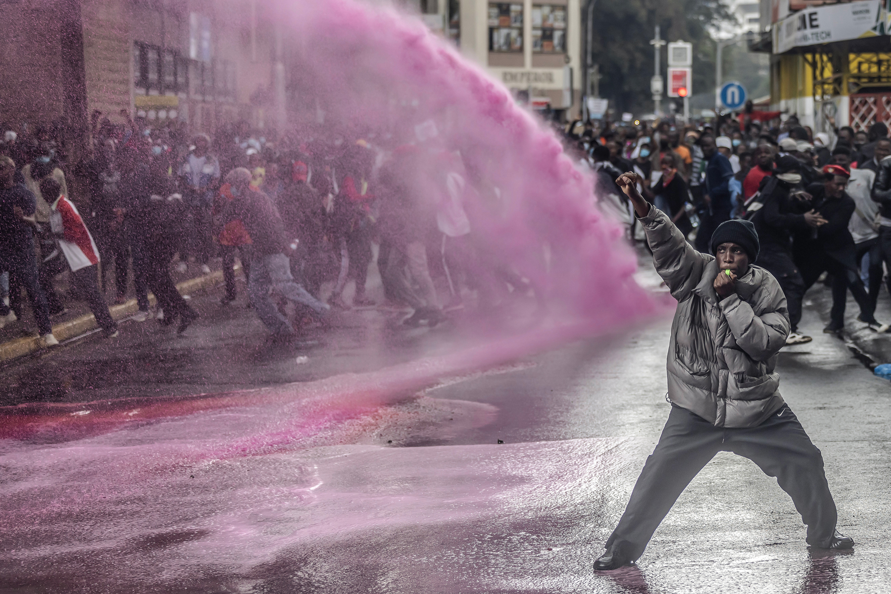 A protestor raises a fist in defiance as Kenyan police officers use a water cannon to disperse the gathering. Pink dye in the water is often used by police at protests to mark individuals for identification. Nairobi, Kenya, 20 June 2024. © Luis Tato, Agence France-Presse