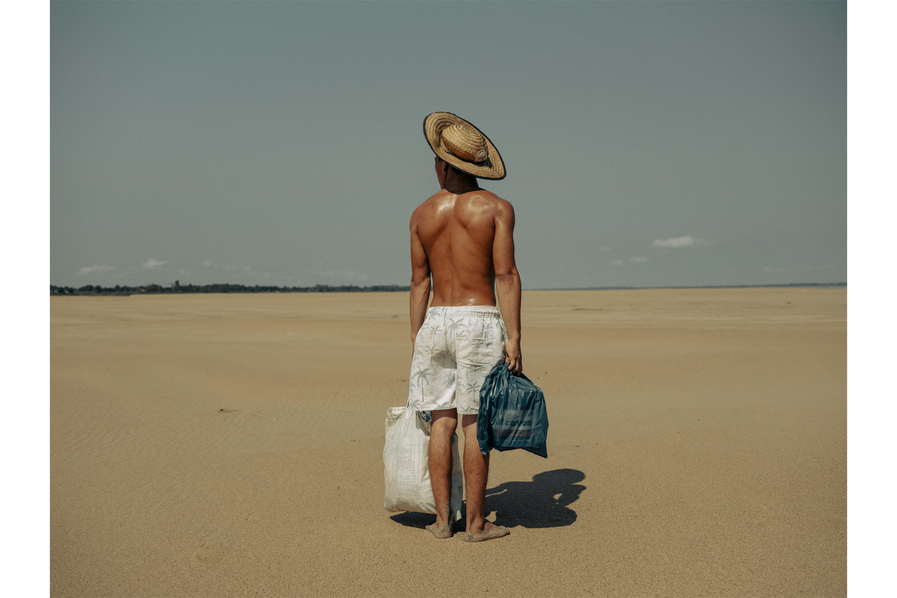 A young man brings food to his mother who lives in the village of Manacapuru. The village was once accessible by boat, but because of the drought, he must walk 2 kilometers along the dry riverbed of the Solimões River to reach her. Amazonas, Brazil, 5 October 2024.    © Musuk Nolte, Panos Pictures, Bertha Foundation